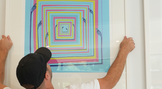 A man in a black cap mounts a large, framed abstract print featuring nested squares and birds in light blue, yellow, and pink on a white wall.