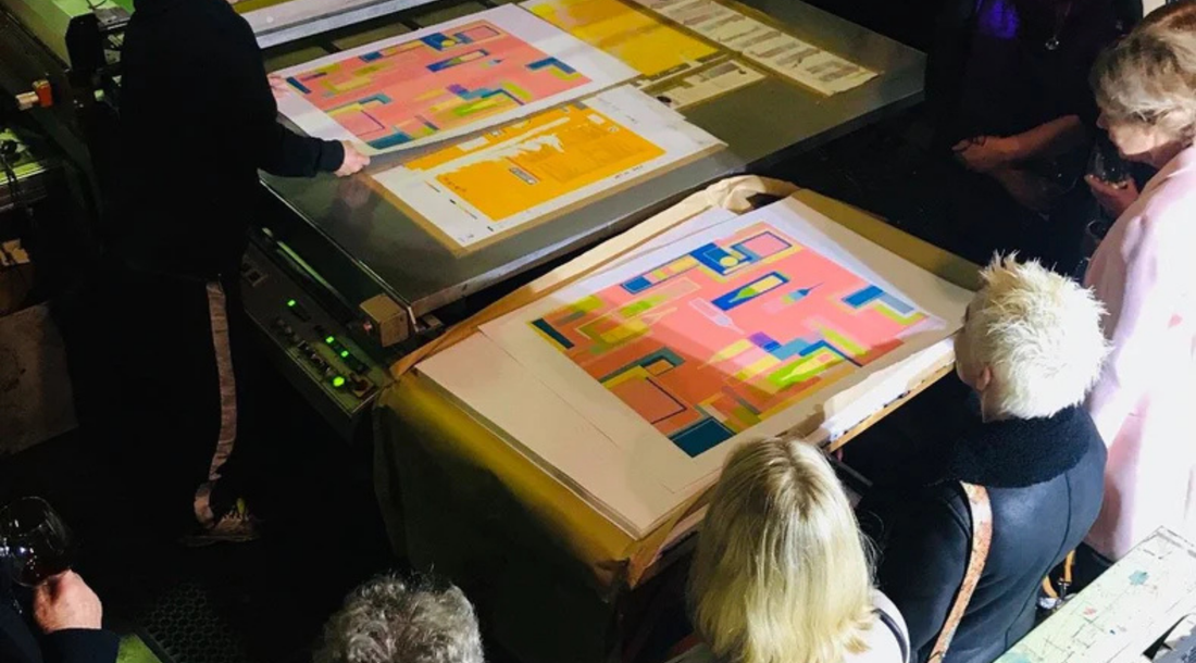 An overhead view of people observing colourful abstract screen prints around a printing press in a studio.