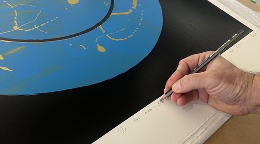 Close-up of an artist's hand signing a blue and black abstract screen print with a pencil, writing the title "In two minds" in the white margin.