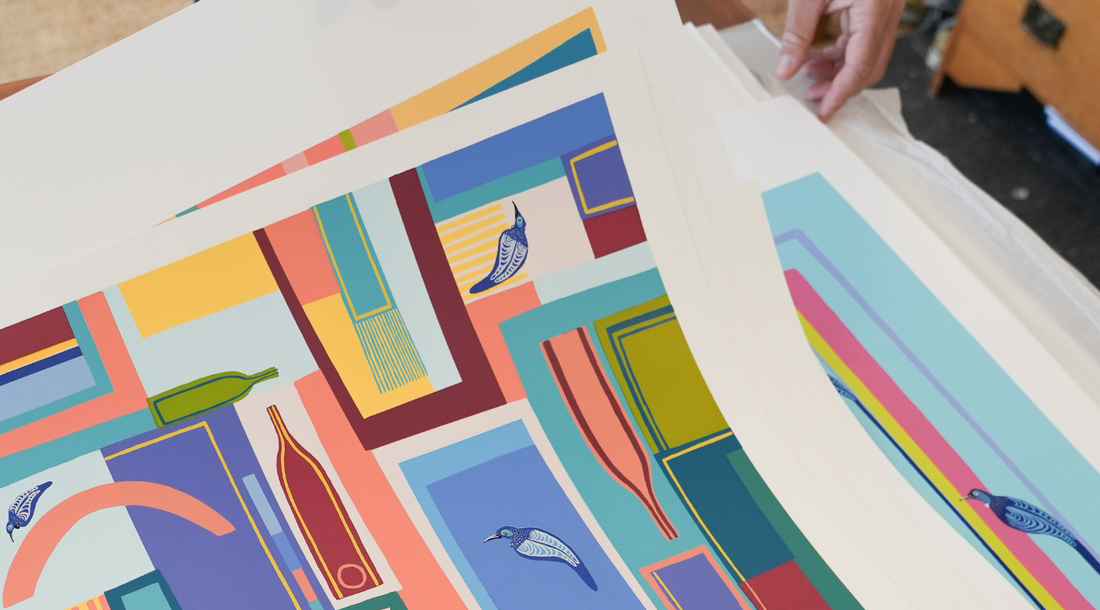 A person's hand sorting through a stack of vibrant, high-quality art prints featuring a modern geometric design with stylized birds and wine bottles in shades of blue, yellow, and red.