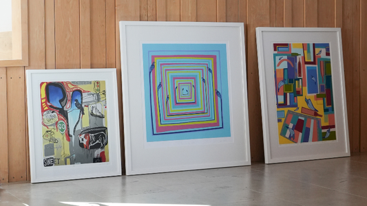 Three large, modern art prints in white frames leaning against a light wood-paneled wall. The colorful illustrations feature geometric patterns, blue birds, and street-art-inspired elements in a vibrant palette of light blue, yellow, and red.