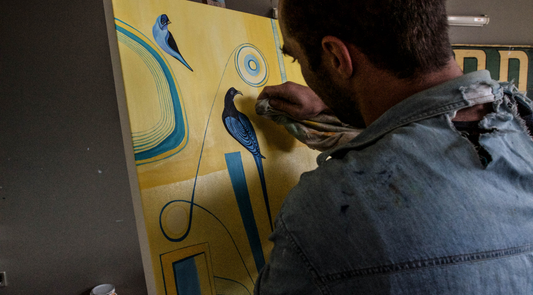 An artist in a worn denim shirt carefully wipes or buffs a bright yellow canvas featuring stylized blue birds and geometric shapes.