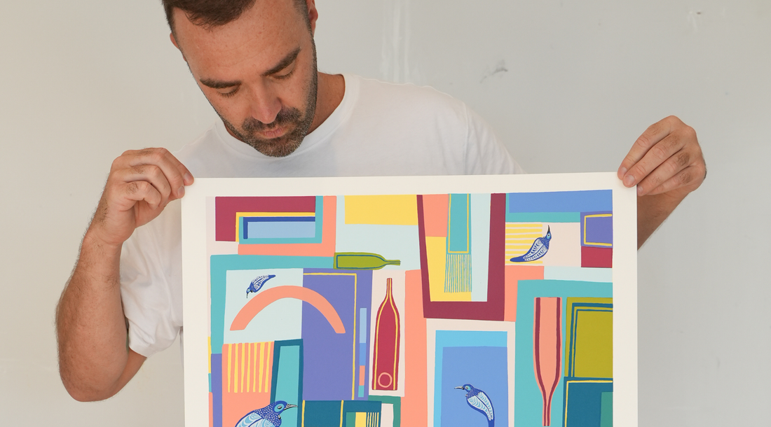 A person in a white t-shirt holding a colorful abstract screen print featuring geometric shapes, bottles, and blue birds.
