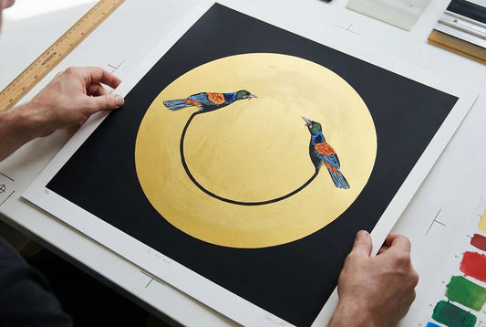 A person’s hands holding a square art print featuring two colorful birds on a black curved line, set against a large gold circle on a black background.